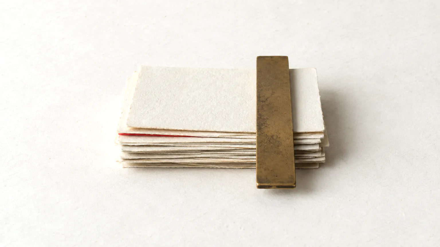 A stack of slightly offset blank paper slips being squared by a plain bronze ruler, one slip edged in red