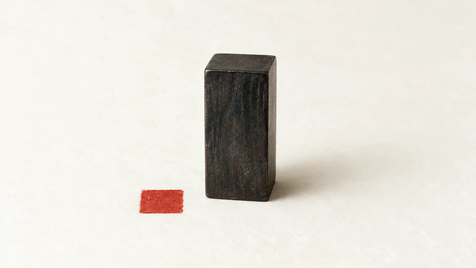 An upright dark wooden seal standing on handmade paper beside a small blank cinnabar square impression