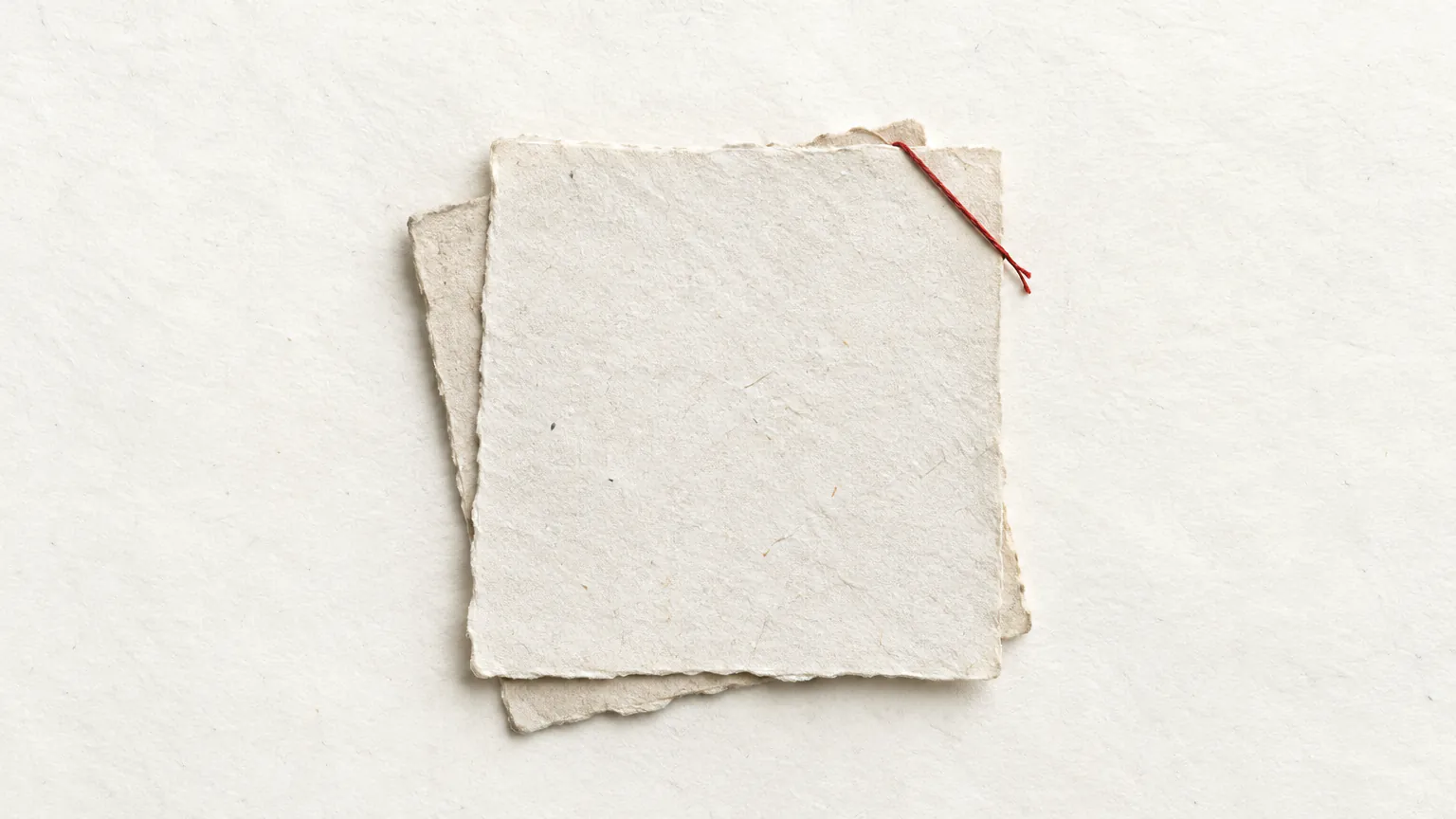 Two blank sheets of handmade paper offset on each other, a red thread across one corner