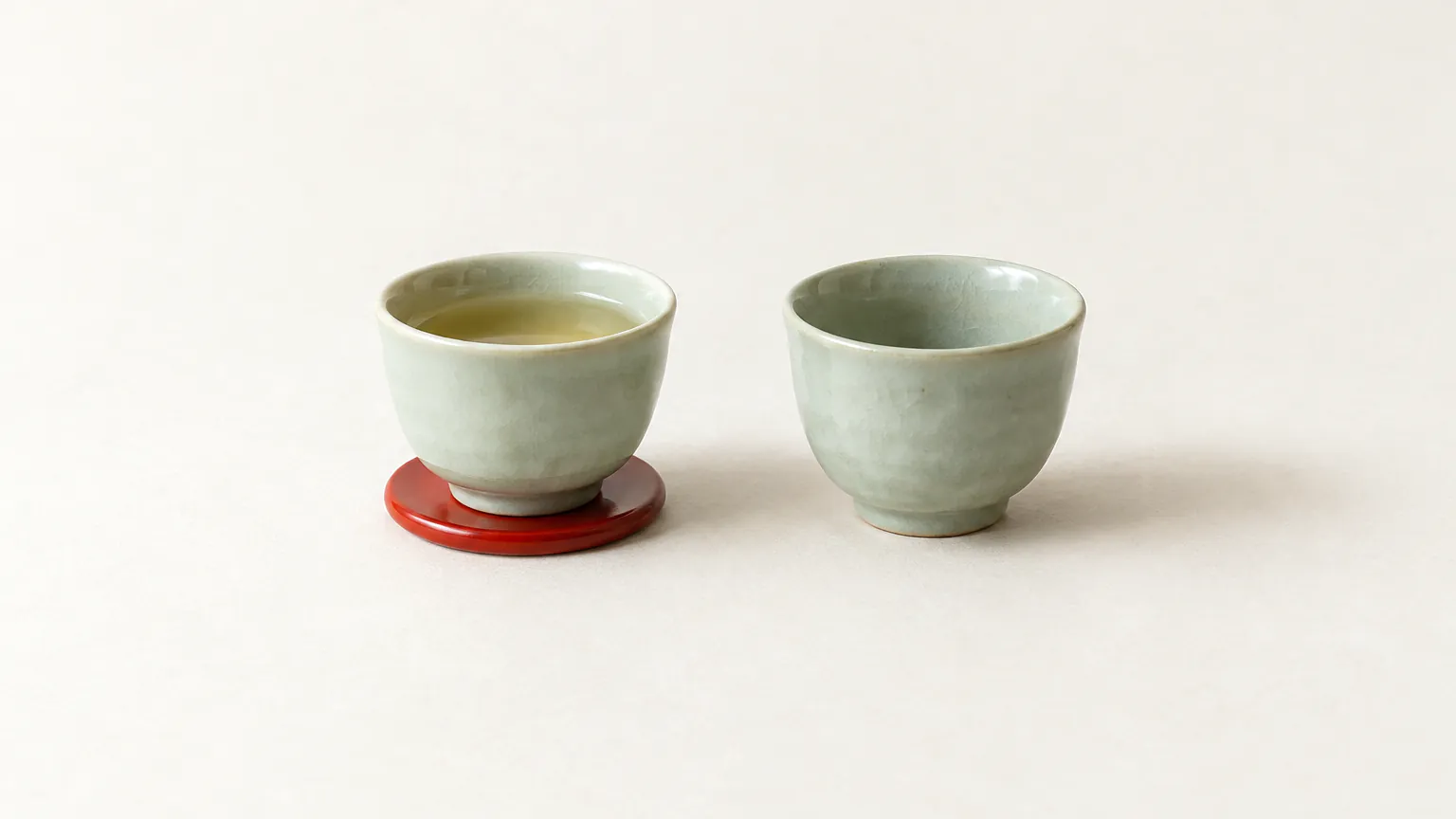 Two small pale celadon tea cups, one half full of pale tea, with a small red lacquer coaster under one cup