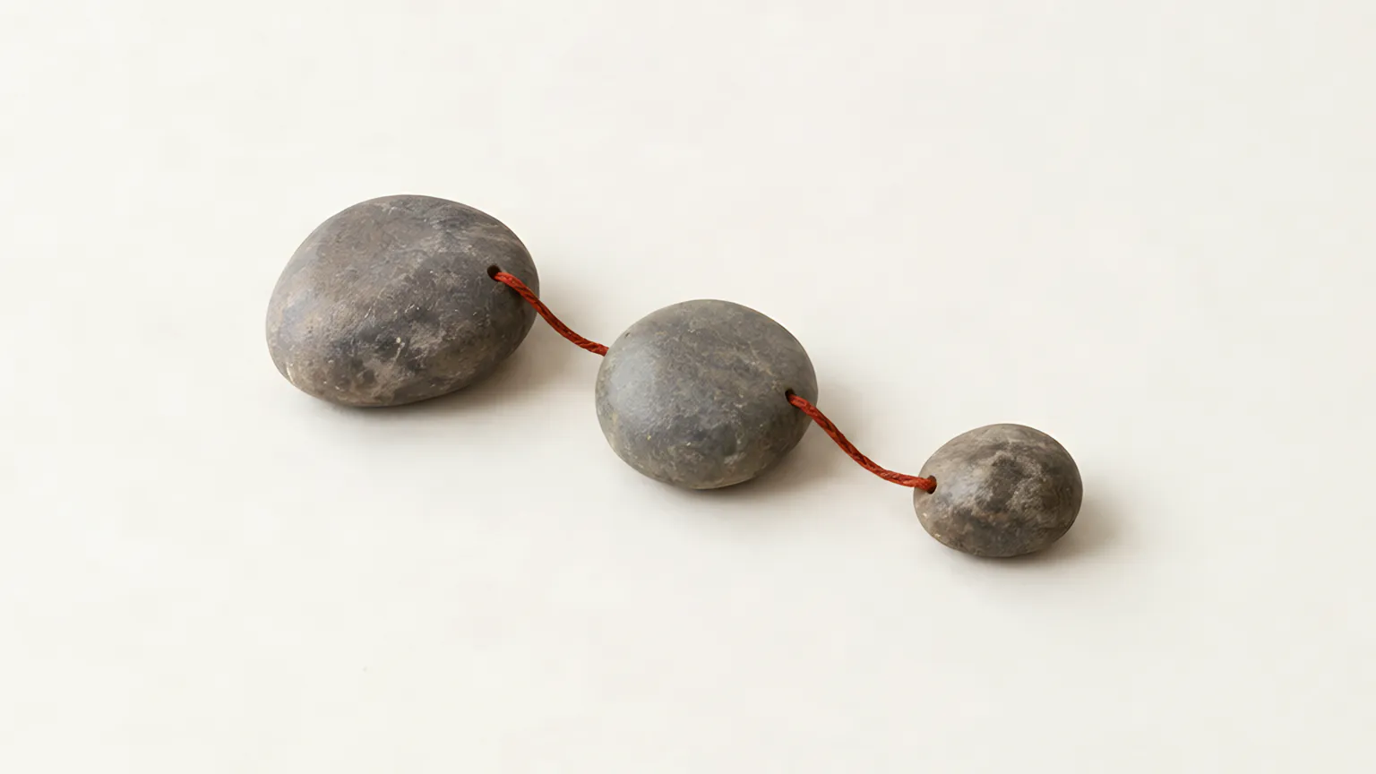 Three grey river stones of decreasing size in a line, joined by a fine cinnabar cord, like a first, middle and final step