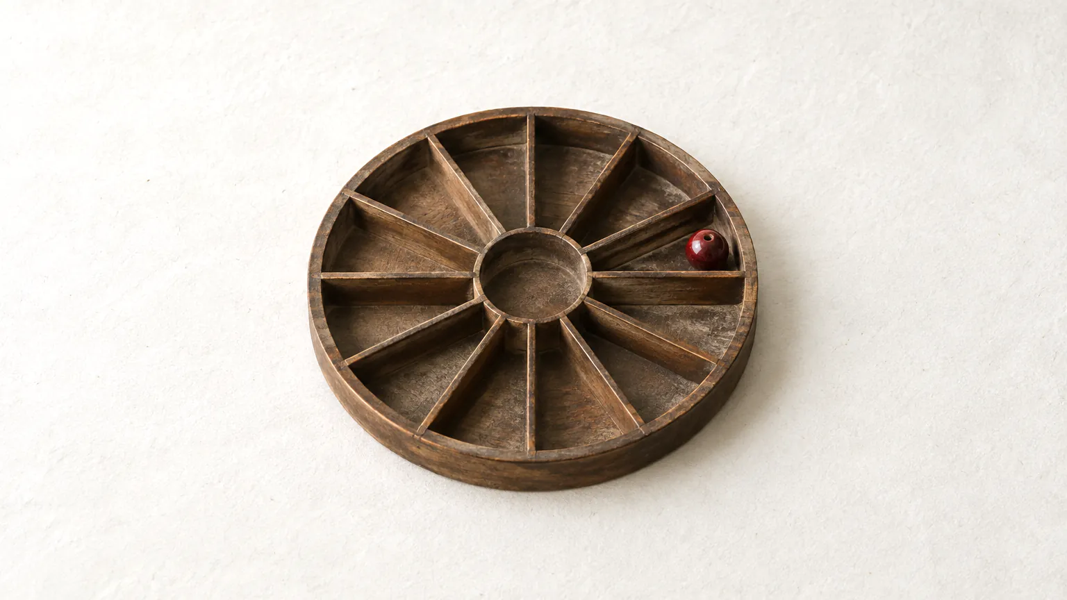 A round wooden tray split into twelve empty compartments, with a single bead in one